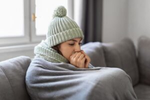 Freezing woman in knitted hat and scarf wrapped in a blanket while sitting on the couch